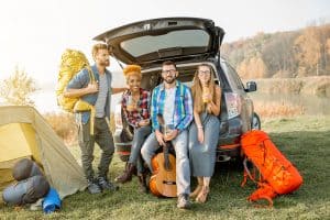 Multi ethnic group of friends sitting at the car trank during the outdoor recreation with tent and hiking equipment near the forest