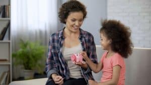 Mother teaching little daughter to save money, throwing coins in piggy bank
