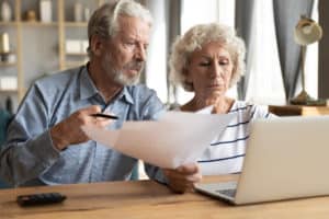 Stressed elderly married couple sitting at table at home manage budget paying using on-line banking access analyzing finance papers, having financial problems, high taxes expenses, unpaid debt concept