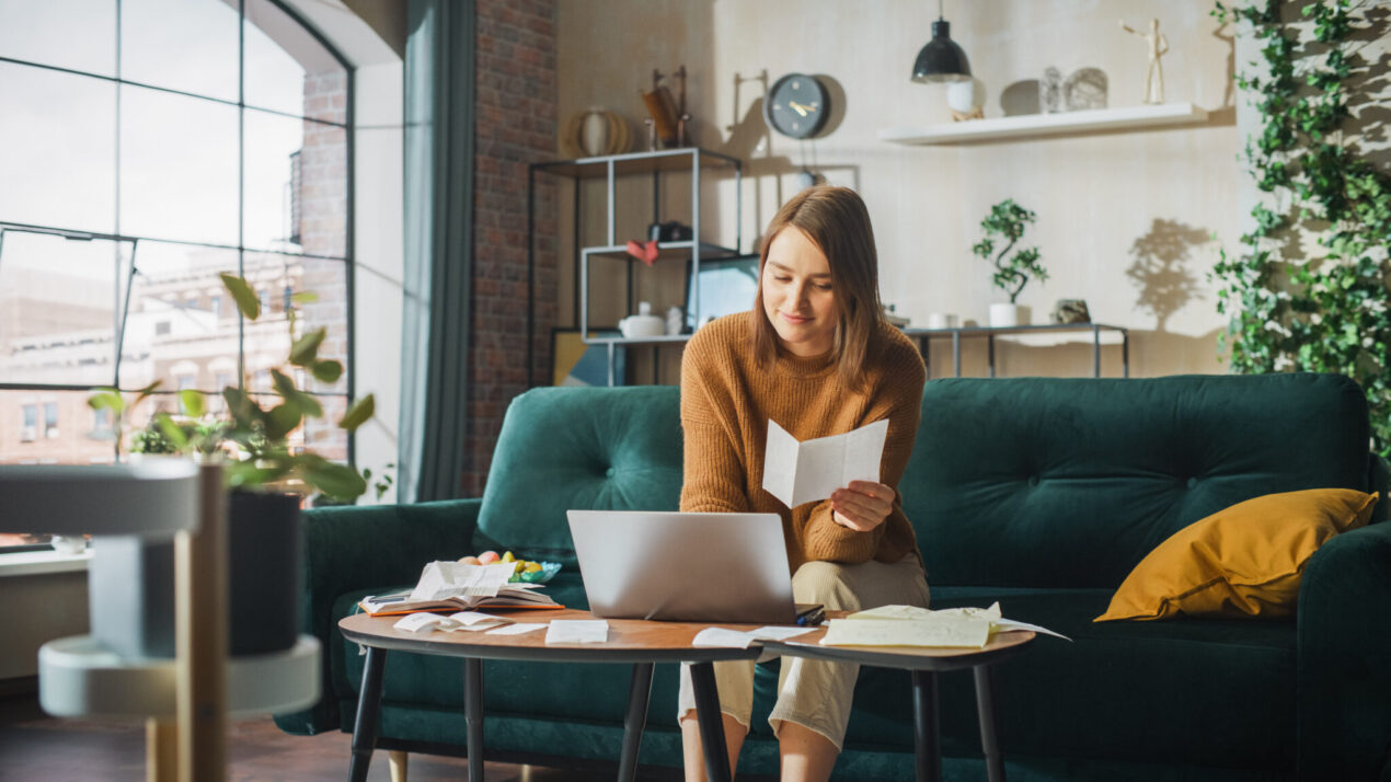 Young woman sitting in an apartment looking at laptop, financial documents