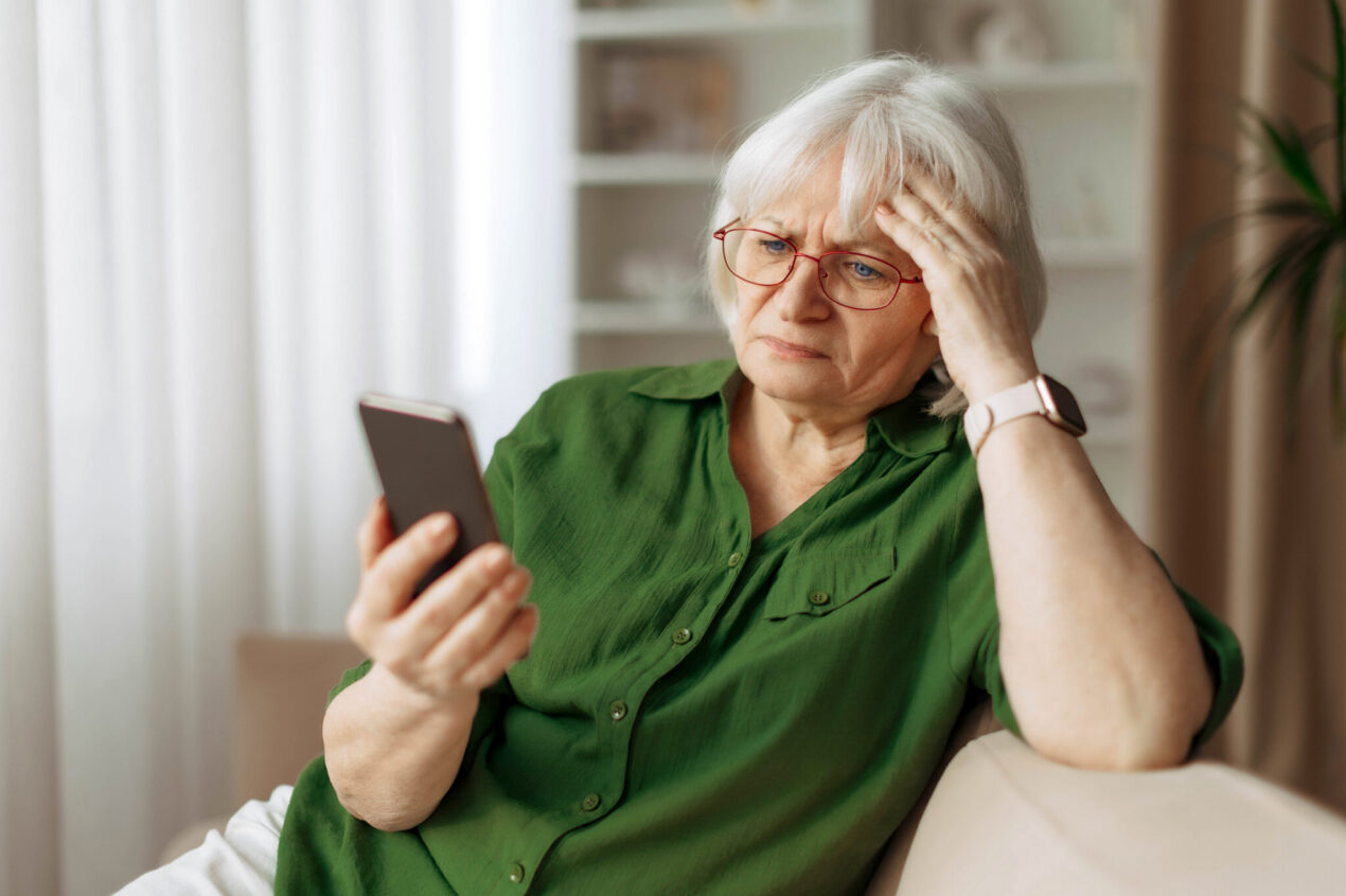Senior woman with glasses looking at smartphone with worried expression while sitting at home. Older person targeted for a scam